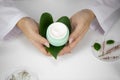 Hands hold jar with cream on green leaf, lab background with herbal ingredients