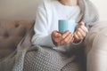 Close up of hands holding blue cup of tea or coffee. Model in white sweater and cozy plaid is sitting, relax at home on sofa Royalty Free Stock Photo