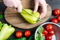 Close up of hands holding a knife and sweet green peppers on an oval wooden chopping Board that lies on a wooden table with