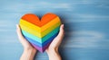 Close-up of hands holding a wood heart mading in the colors of the LGBT flag on blue background. Love and Relationships concept.