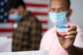 Close up of Hands, Man in medical mask showing I voted Sticker at polling booth with US flag as background - concept in person