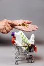 Close up of hands of senior woman counting a coinson near full of medicines little shopping cart. Gray background