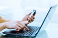 Close-up of hands typing on laptop computer while using a cell phone.