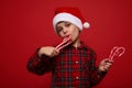 Close-up of handsome adorable boy in plaid shirt and Santa Claus hat tasting sweet sugary Christmas lollipops, striped candy canes