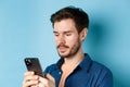 Close up of handsome bearded man reading mobile phone message, looking at screen serious, standing on blue background