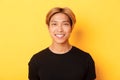 Close-up of handsome blond asian guy in black t-shirt, smiling happy at camera, standing over yellow background