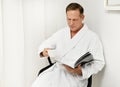 Close-up of a handsome European mature business man wearing white terry bathrobe, sitting on an armchair and reading magazine Royalty Free Stock Photo