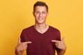 Close up of handsome man in blank burgundy t hirt pointing at himself isolated over yellow studio background, male with pleasant