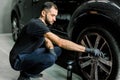 Close up of handsome young male employee worker cleaning the wheel rim of modern black crossover car by scrubbing brush