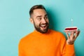 Close-up of happy adult man celebrating birthday, holding bday cake with candle and making wish, standing against