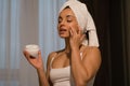 Close up of happy Caucasian beautiful young woman after shower with towel on head using moisturizing face cream at home Royalty Free Stock Photo