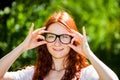 Close-up of a happy red-haired woman adjusting her glasses in a large black frame, smiling in nature with bokeh. Royalty Free Stock Photo