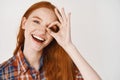 Close-up of happy redhead woman with pale skin and white teeth, showing okay sign on eye and smiling, standing against