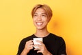 Close-up of happy smiling handsome blond asian guy, looking away with dreamy nostalgic look while drinking coffee