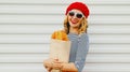 Close up of happy smiling young woman wearing a french red beret holding paper bag with long white bread baguette over a white
