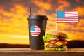Close-up home made beef burger with american flag on the top and cup on wooden table over sunset background Royalty Free Stock Photo