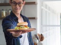 Close-up of a homemade hamburger on a wooden dish, holding by young man stand wearing glasses looking at the camera, smiling. Royalty Free Stock Photo