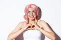 Close-up of hopeful smiling girl in pink party wig, looking up and showing heart sign, standing over white background Royalty Free Stock Photo