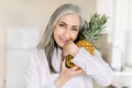 Close up horizontal beauty shot of happy senior gray haired woman holding fresh pineapple, looking at camera with smile