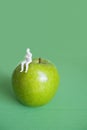 Close-up of human figurine sitting on green apple over colored background