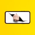 Close-up of human fist punching through white paper hole on screen of smartphone; isolated on background of yellow color. Royalty Free Stock Photo