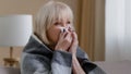 Close up ill sick old blonde grandmother 60s woman covered in warm blanket at home blowing nose to paper napkin suffers Royalty Free Stock Photo