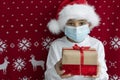 Close up image of boy in santa hat and face medical mask holds two gift boxes tired with ribbons on red christmas background.