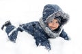 Close-up image of cute baby boy in a warm white snow suit lying in snow with funny face.