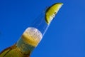 Close up of isolated bottleneck with sparkling yellow beer and a slice of lime against cloudless deep blue sky