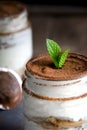 Close up of a jar filled with tiramisu with cocoa powder on top and mint leaves as decoration and a second vase on the background
