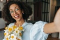 Close-up of joyful young african woman taking selfie with bouquet of daffodils at home.