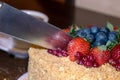 Close-up kitchen knife cutting of cake decorated with mixed berries, settled on wooden table. Royalty Free Stock Photo