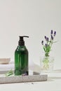 Close-up of a labelless transparent green shampoo bottle displayed on a gray tray, placed next to a vase of lavender flowers,