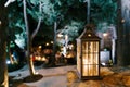 Close-up of a lantern candlestick with a candle inside on a stone texture in a courtyard with trees and people.