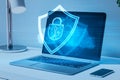 Close up of laptop, cellphone and lamp with glowing shield hologram on screen. Blurry desktop background. Secure, protection,