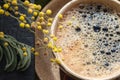 Close-up on a large foam in a ceramic cup of fragrant morning coffee. Nearby are a part of a reading book on which yellow mimosa