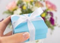 Little gift box with white ribbon in female hand in the background of the bouquet of flowers Royalty Free Stock Photo