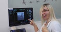 Close-up looking at the camera. A beautiful blonde doctor funny shows her thumbs up and points to the monitor of the