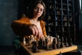 Close-up low-angle view of thoughtful young woman in elegant eyeglasses making chess move sitting in armchair in dark Royalty Free Stock Photo