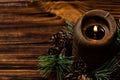 A lit brown candle is decorated with a spruce branch with small cones. Brown wooden boards on the background.