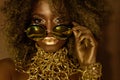 Close-up of magic golden african american female model in massive sunglasses with bright glitter makeup, glossy