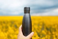 Close-up of male hand holding black steel thermo water bottle on background of blurred rapeseed field. Royalty Free Stock Photo