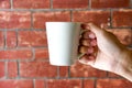 Close up of male hand holding blank white coffee or tea cup on old red brick wall background texture with morning light Royalty Free Stock Photo