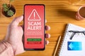 Close up of male hand holding mobile phone with red screen and warning icon with scam alert text and message saying that the site