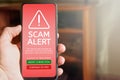 Close up of male hand holding mobile phone with red screen and warning icon with scam alert text and message saying that the site