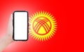 Close-up of male hand holding smartphone with blank on screen, on background of blurred flag of Kyrgyzstan