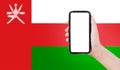 Close-up of male hand holding smartphone with blank on screen, on background of blurred flag of Oman