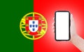 Close-up of male hand holding smartphone with blank on screen, on background of blurred flag of Portugal.