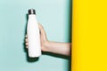 Close-up of male hand holding steel reusable thermo water bottle of white with mockup, between two backgrounds. Royalty Free Stock Photo