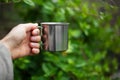 Close-up of male hand holding steel thermo mug on background of green bush in bokeh.